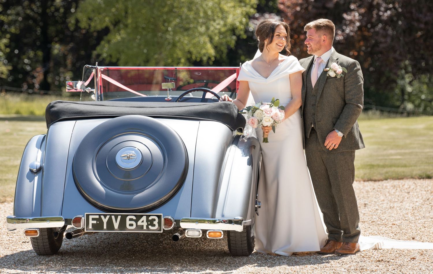 Newlyweds pose beside classic silver vintage car at countryside venue.