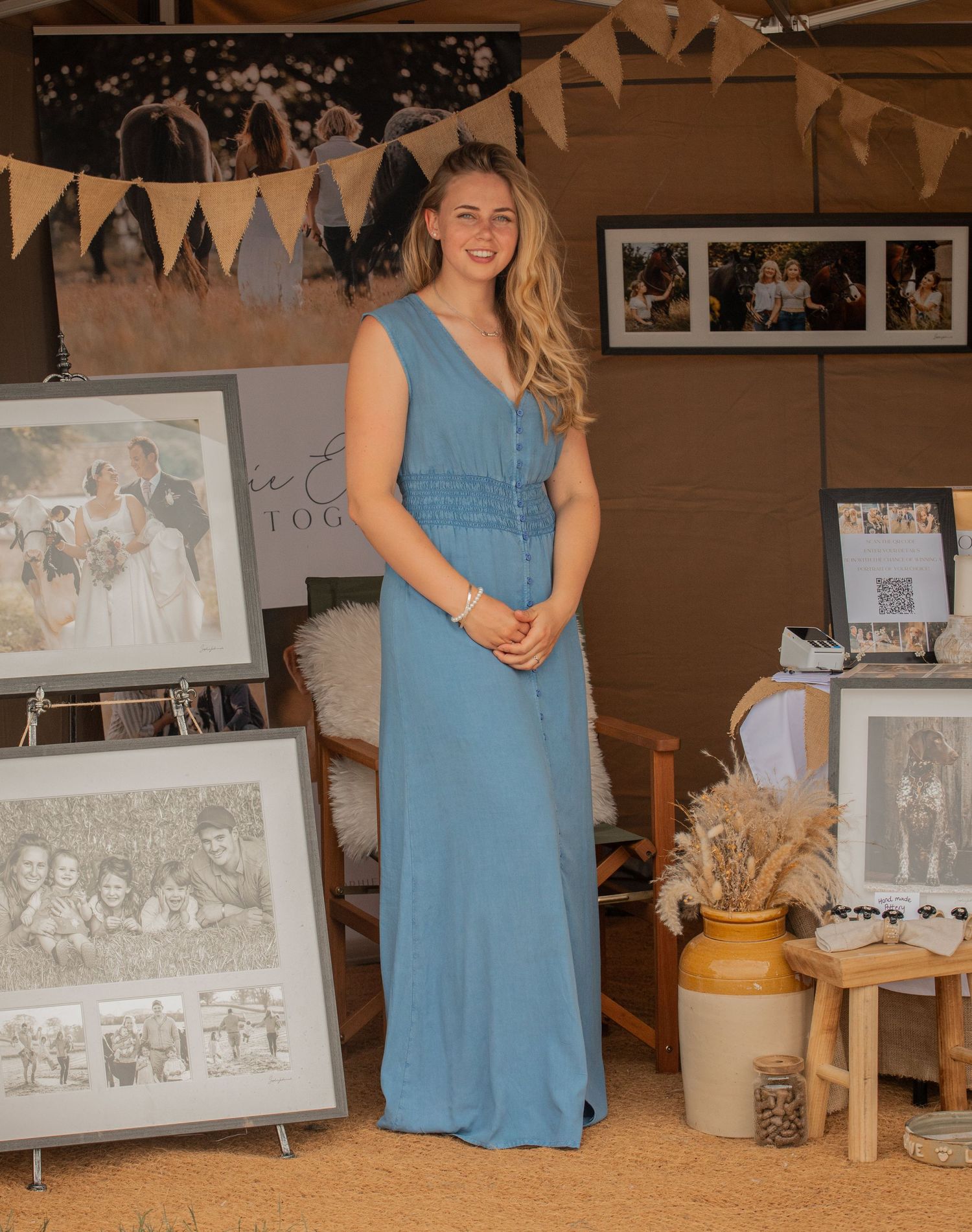 Woman in blue maxi dress stands at a photography booth displaying framed wedding and family portrait prints.
