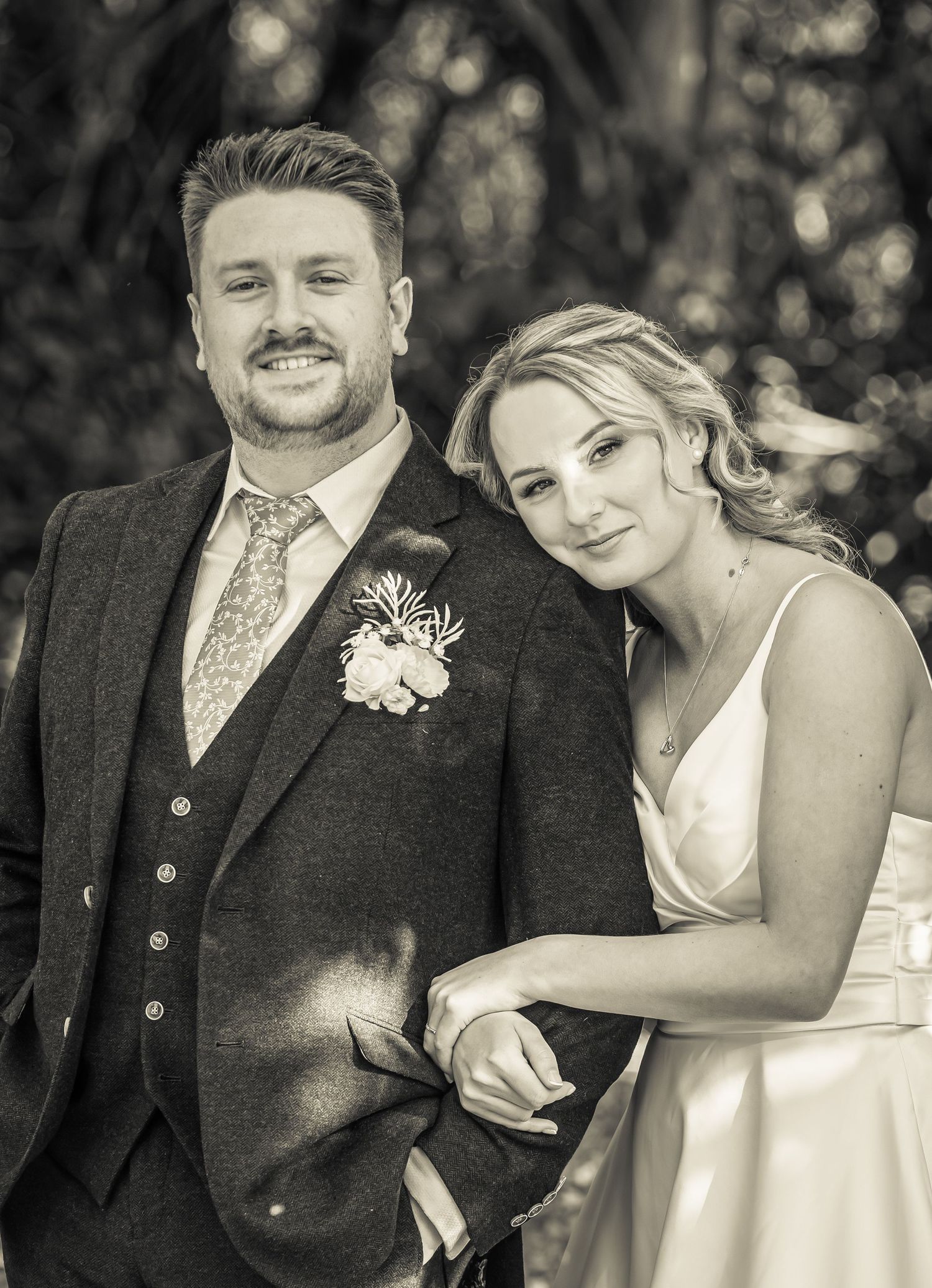 Black and white wedding portrait of smiling groom in tweed suit with boutonniere beside bride in white dress.