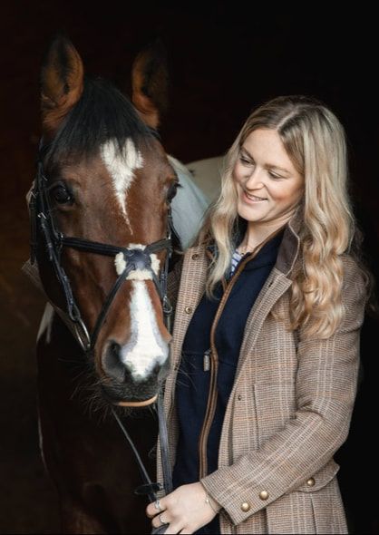 A woman in a brown plaid coat and navy top shares a tender moment with a brown horse with white markings in a dark setting.