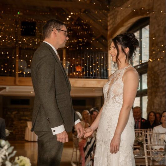 A couple holds hands during their wedding ceremony under twinkling lights in a rustic indoor venue.