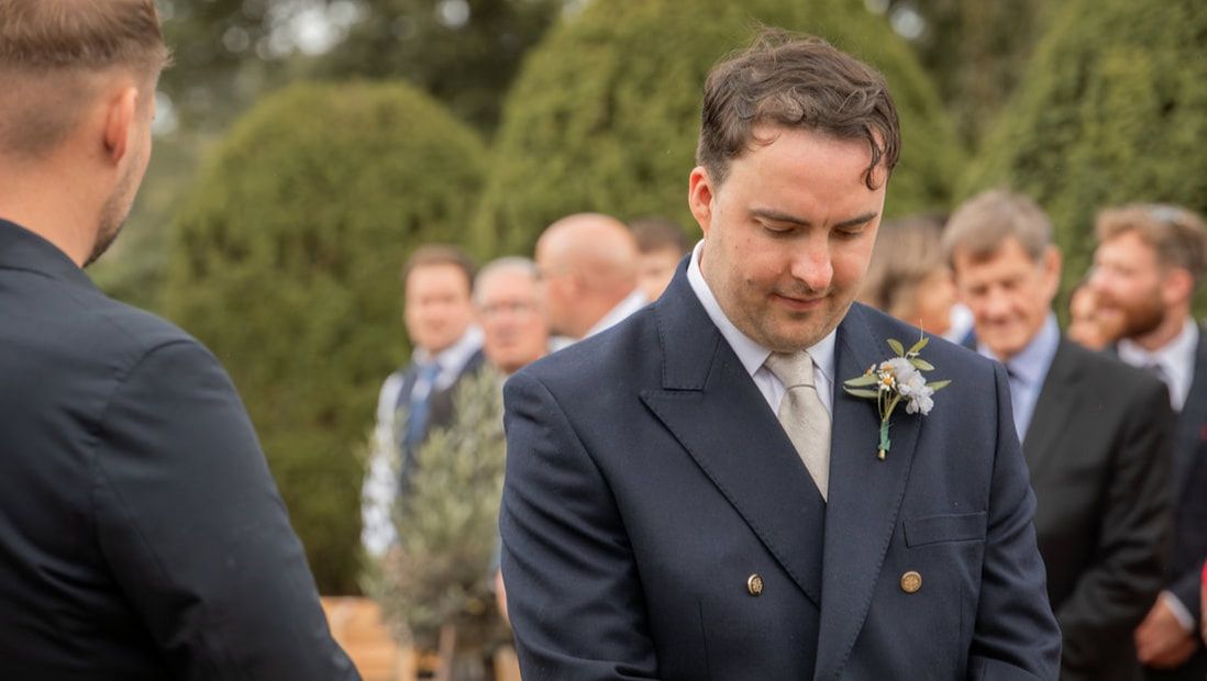 A wedding ceremony takes place outdoors with guests gathered under green trees on a sunny day.