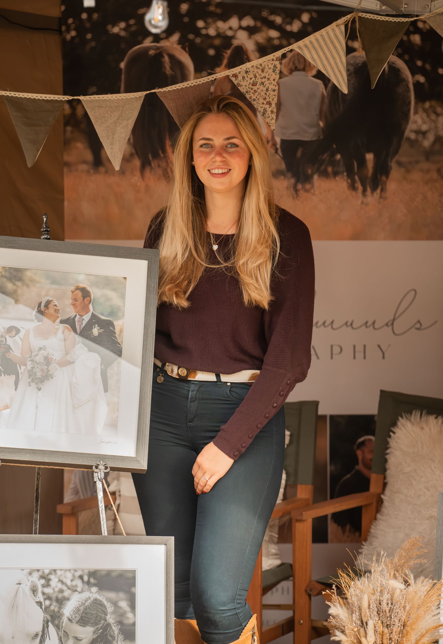 Smiling woman with long blonde hair standing beside wedding photos at a photography exhibition booth.