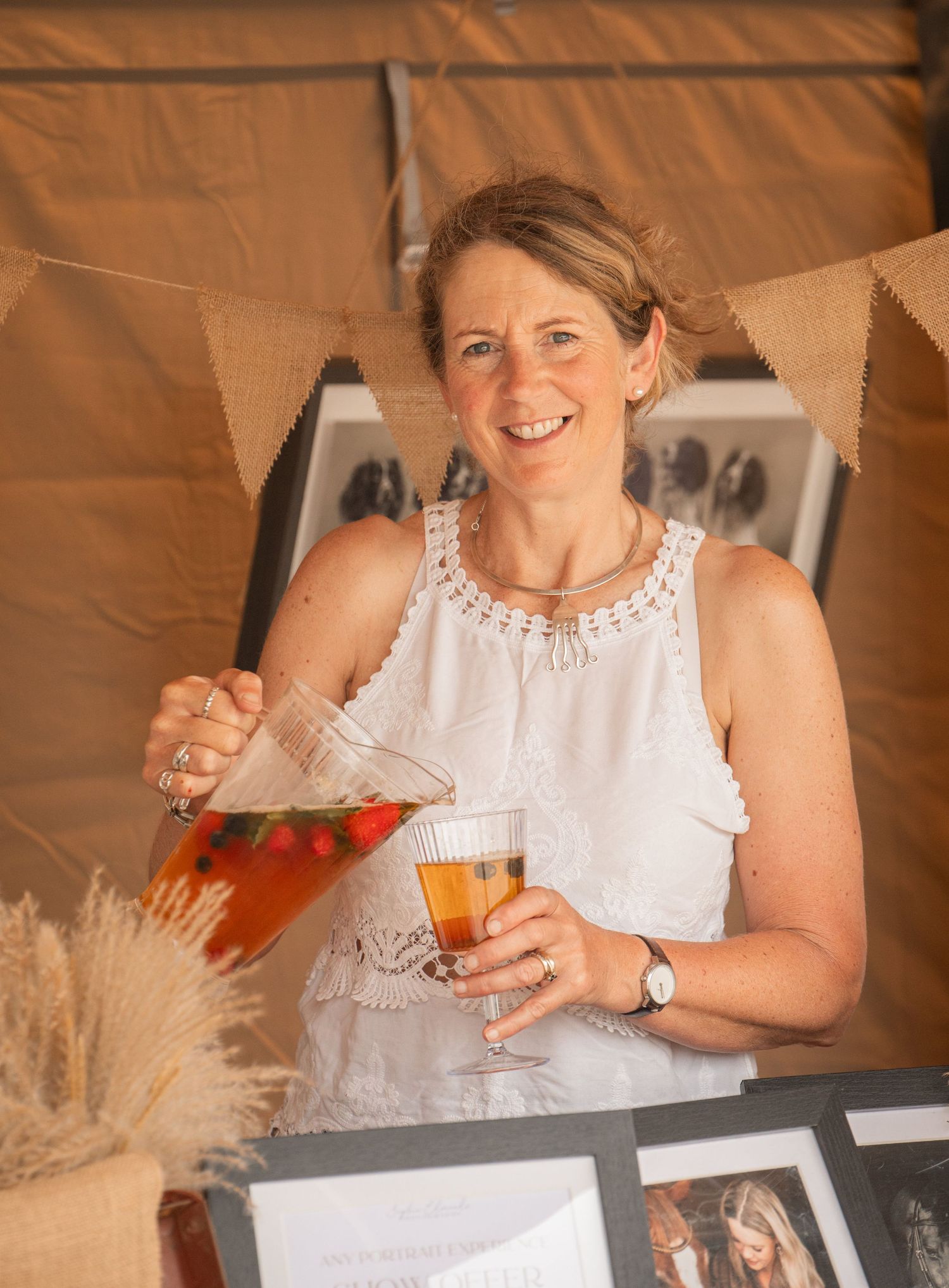 Woman in white dress pouring fruit-infused drink from glass pitcher at outdoor event with bunting decorations.