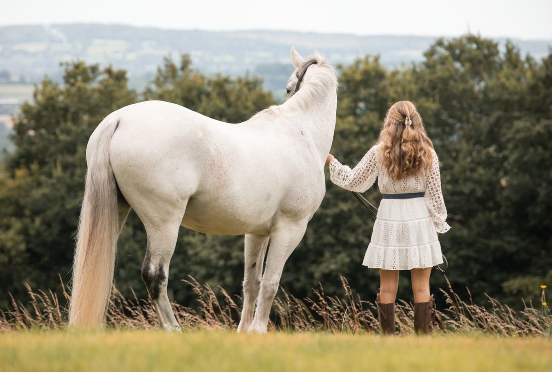 White horse and person in a white dress stand together in a grassy field with trees in the background.