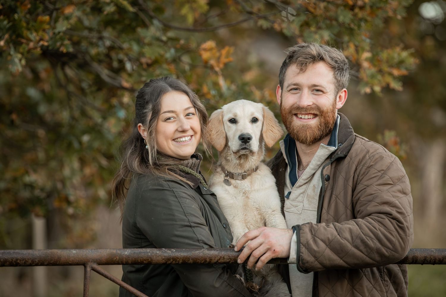 A couple poses with their golden retriever dog by a rustic wooden fence during autumn.