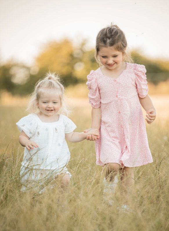 Two children in white and pink dresses walk together through a golden field at sunset.