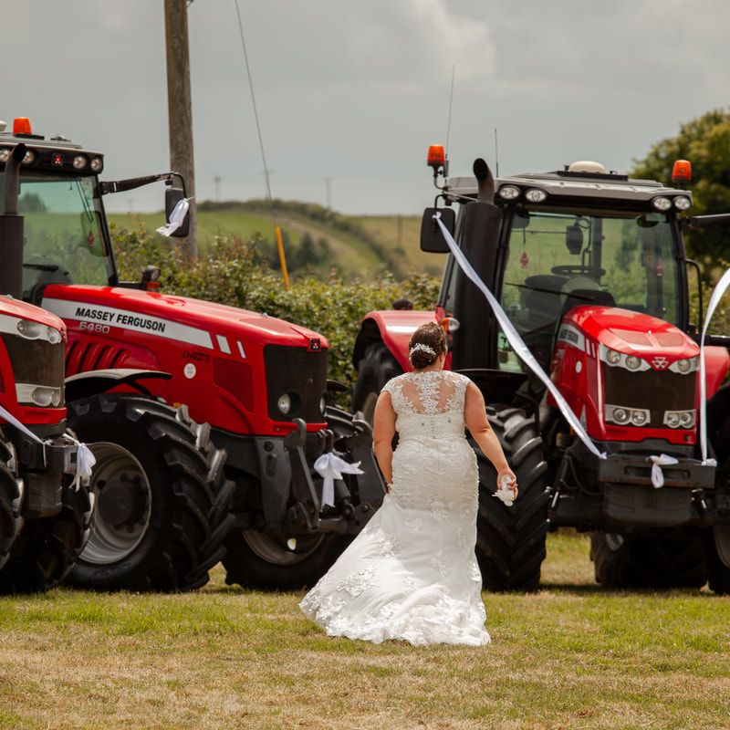A row of red tractors and farm equipment lined up outdoors with a bride in a white wedding dress posing among them.