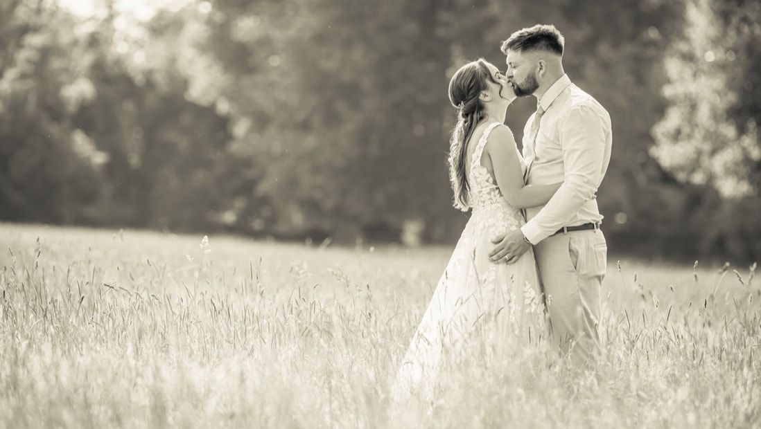 A romantic couple in light-colored attire embraces in a dreamy outdoor garden setting with soft natural lighting.