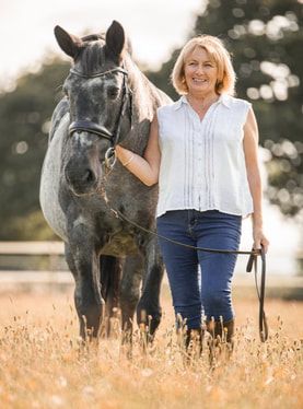 A woman in a white shirt stands beside a large black horse in a golden wheat field at sunset.