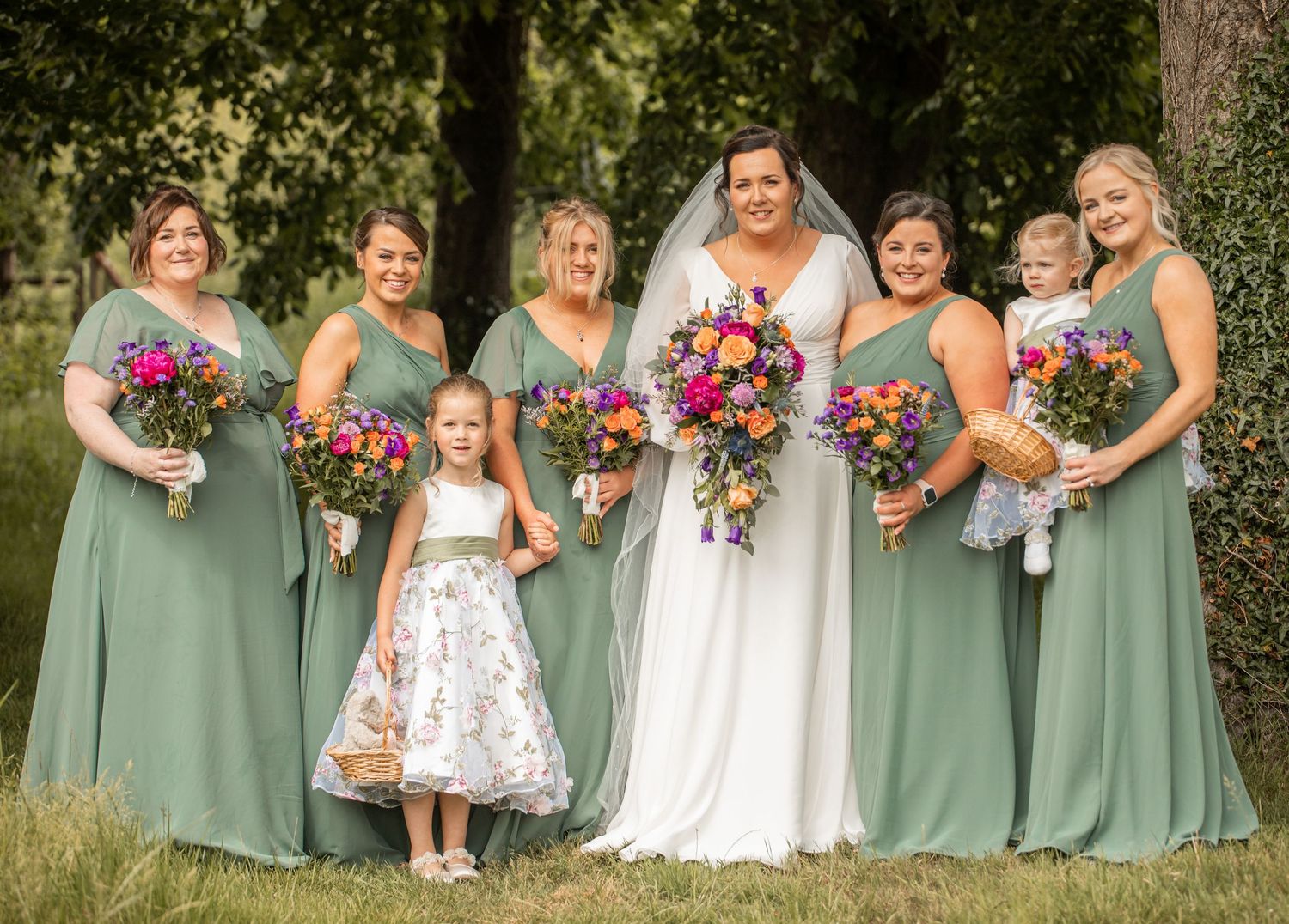 Wedding party wearing sage green bridesmaid dresses holding vibrant floral bouquets pose together outdoors.