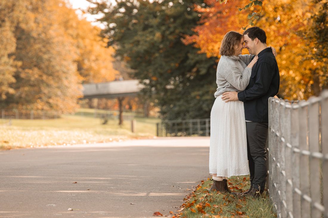 A couple shares a romantic moment on a bridge surrounded by vibrant autumn foliage in a park setting.