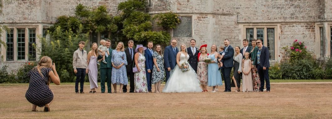 A panoramic view of wedding guests gathering outside a historic stone building with arched windows.