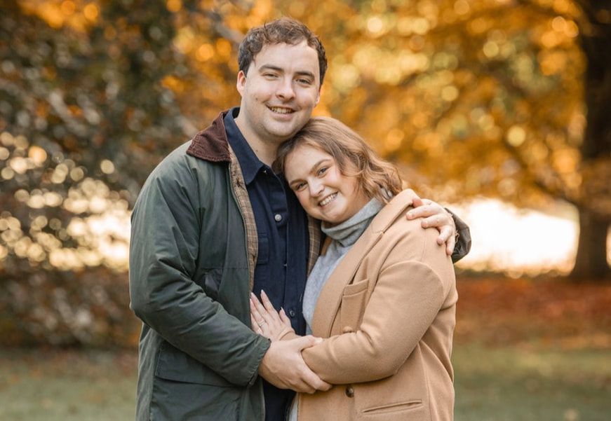 Couple embraces outdoors during golden hour autumn photoshoot with warm fall foliage in background.