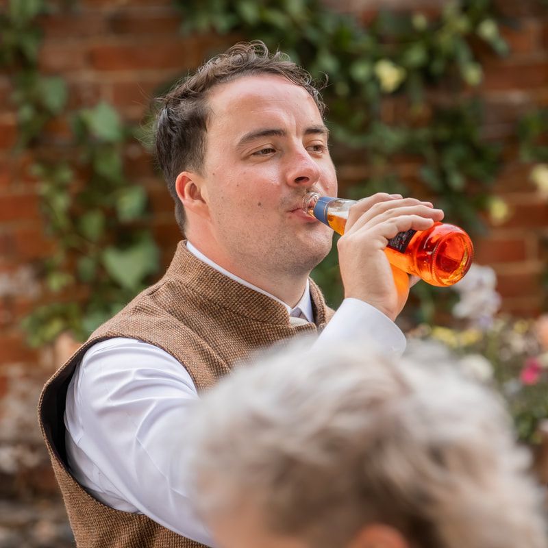 A person in white shirt and brown vest enjoys a drink outdoors against a brick wall with greenery.