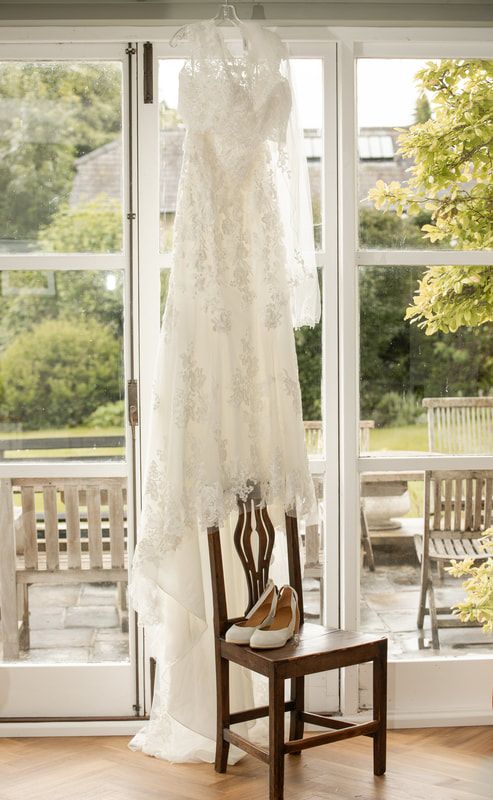 A white lace wedding dress hangs elegantly in front of a sunlit window with a wooden bench in the foreground.