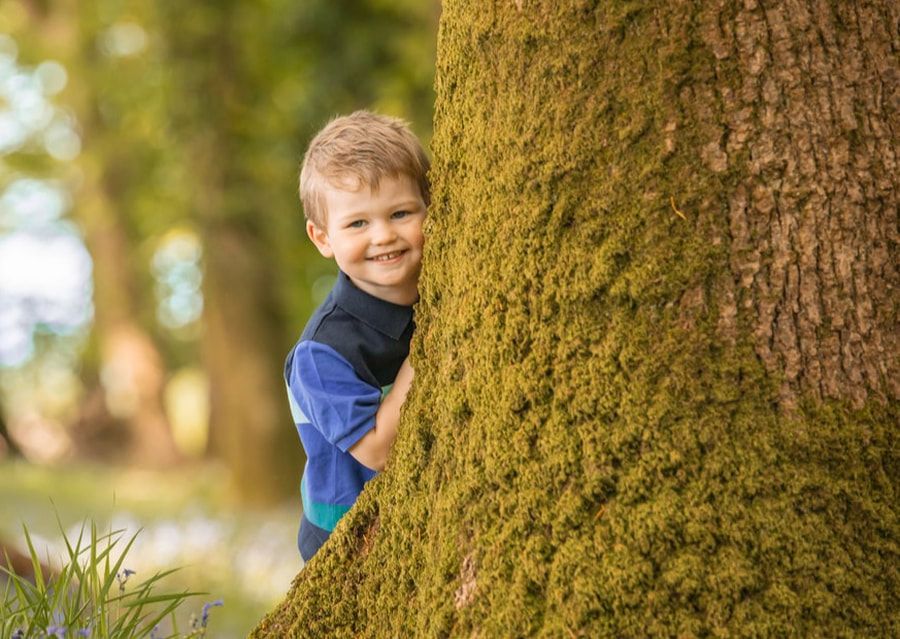 A young child peeks playfully from behind a large mossy tree trunk in a sunlit outdoor setting.