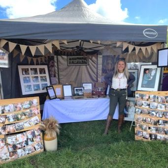 Outdoor craft fair booth displays with white tents and photo prints arranged on tables under a blue summer sky.
