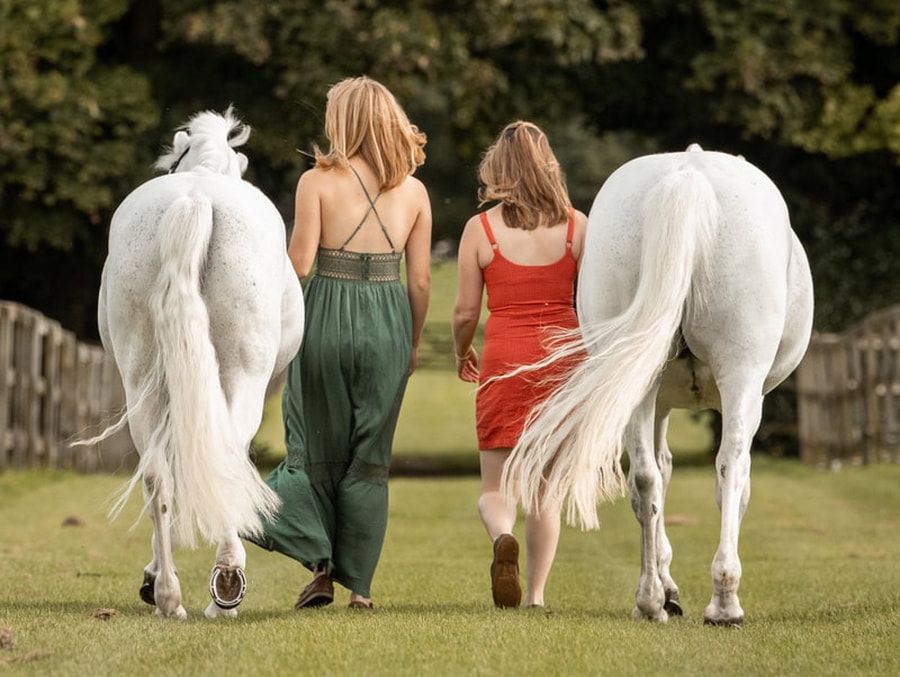 People walking alongside white horses on grassy field during outdoor photoshoot.
