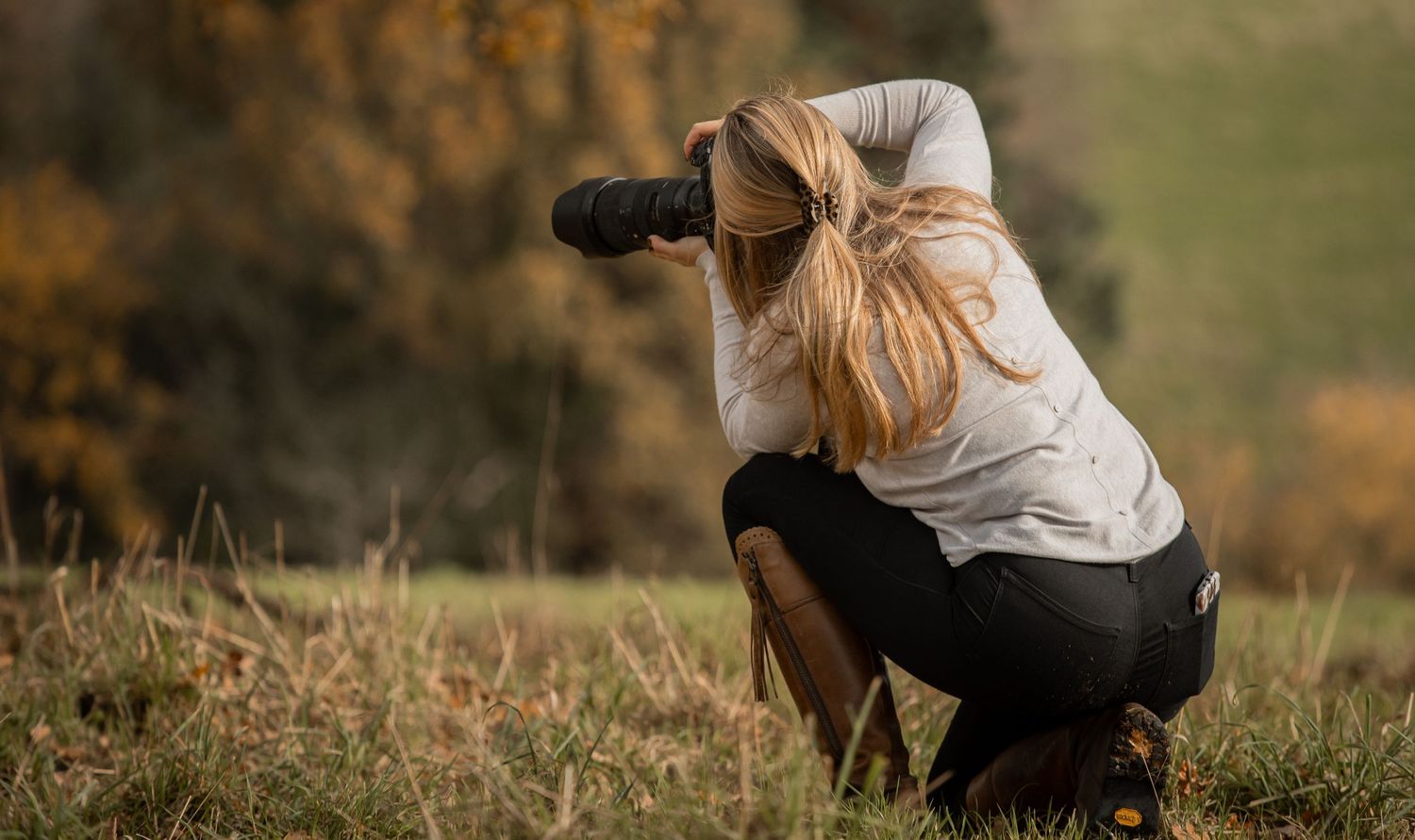 Professional photographer crouching with camera in a grassy field during golden hour.