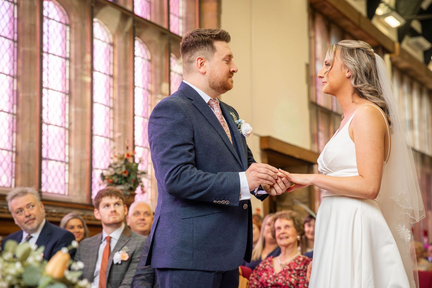Bride and groom exchange rings at wedding ceremony in elegant church with stained glass windows and seated guests.