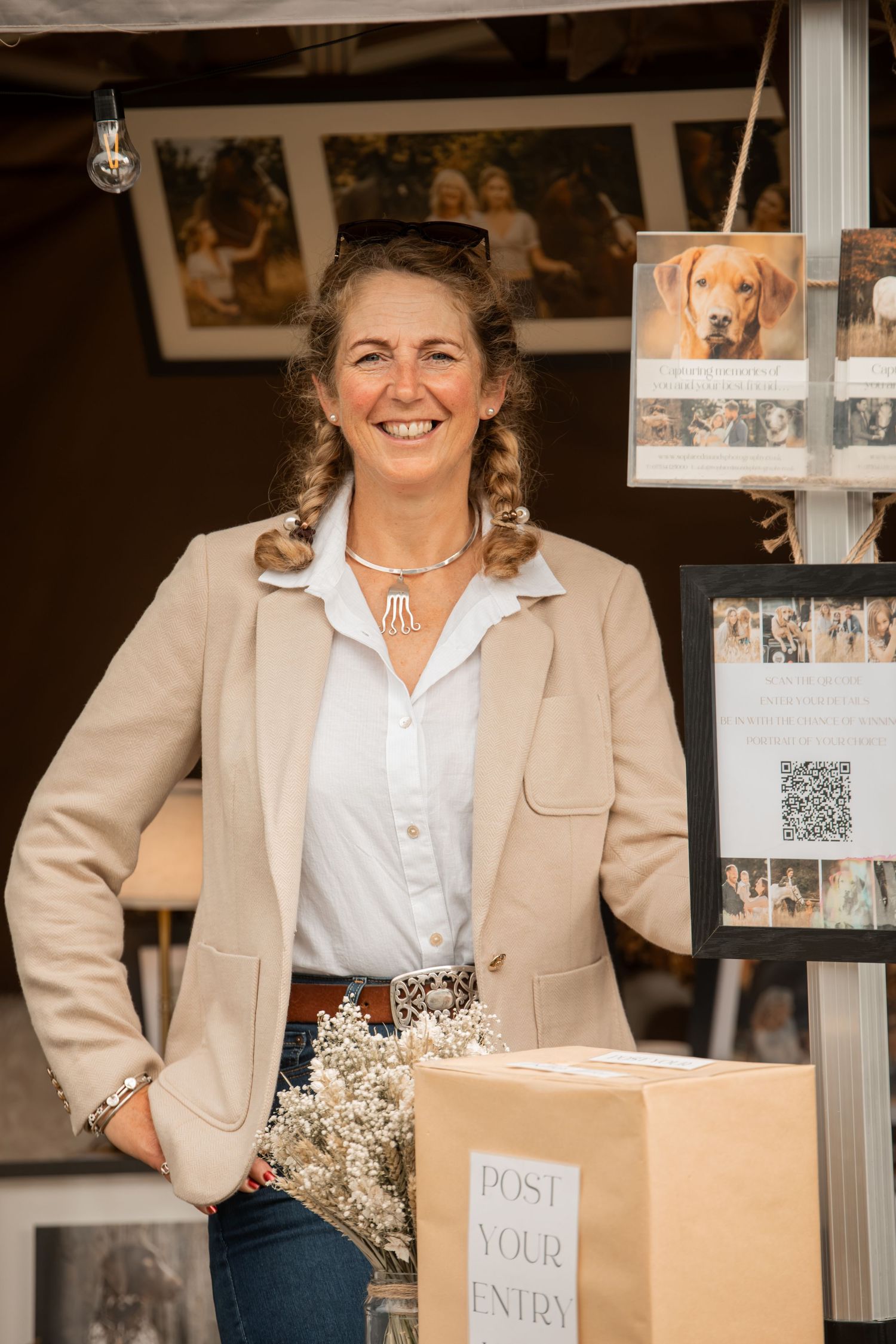 Smiling woman in beige blazer stands at a craft fair booth with a post your entry box and dog photos on display.