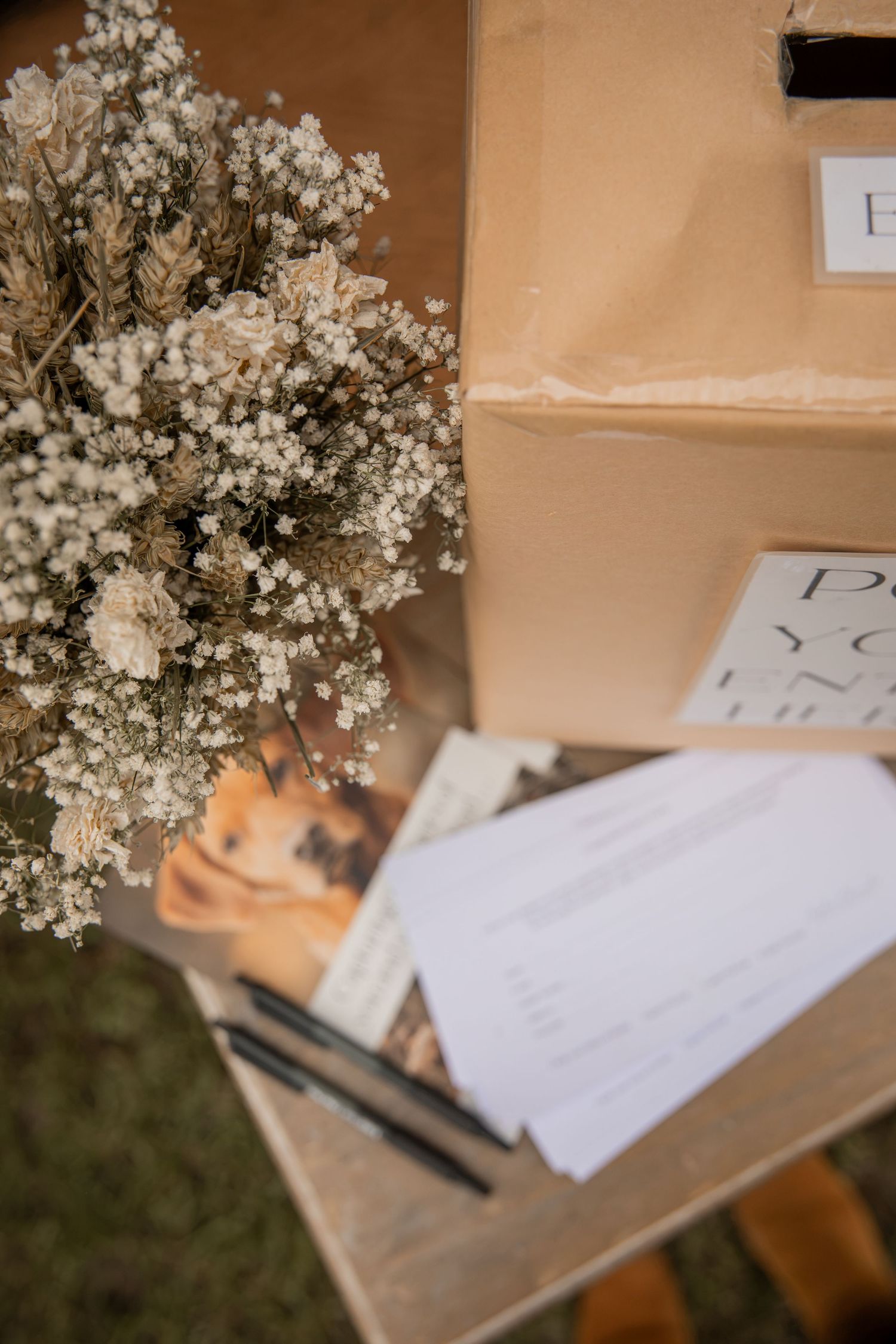 Rustic wedding card box with dried baby's breath flowers, pens, and guest cards on a wooden table.