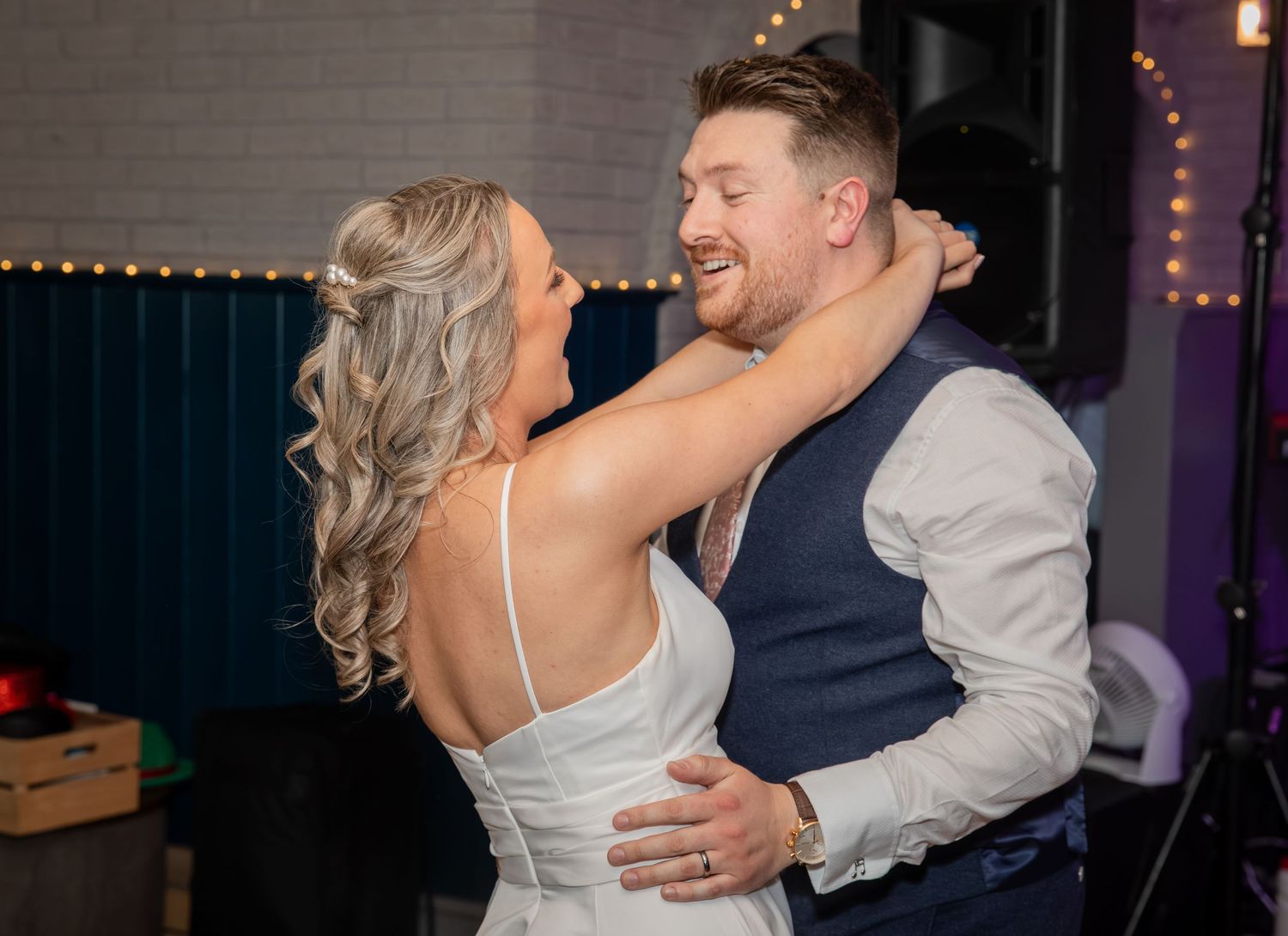 Bride and groom sharing a joyful first dance at their wedding reception with warm string lights in background.