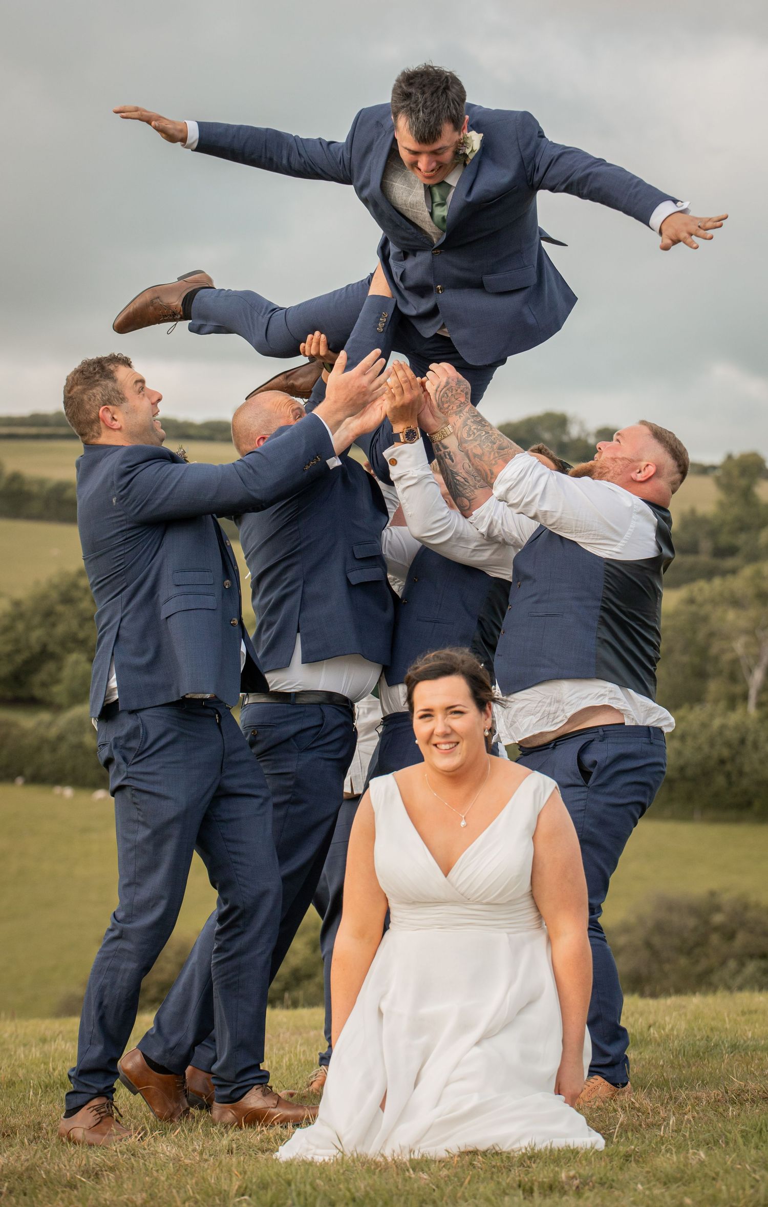 A fun wedding party poses for creative action shots in navy suits and white dress on a grassy hillside.