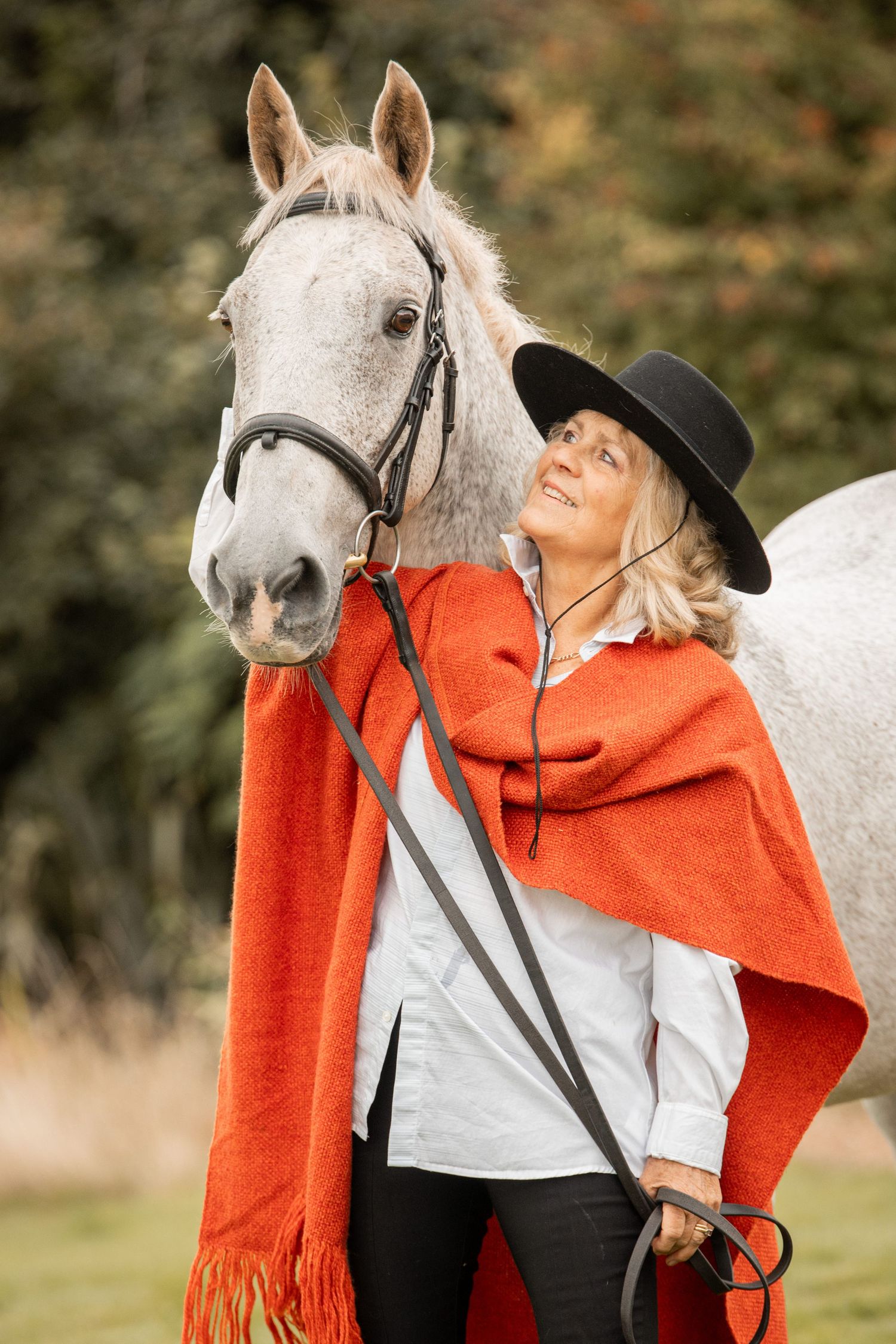 Woman in orange scarf and black hat showing affection to a white horse in an outdoor setting.