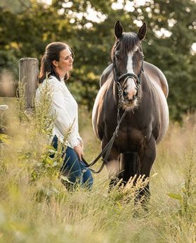 A black horse and person stand in a tall grass field during summer.