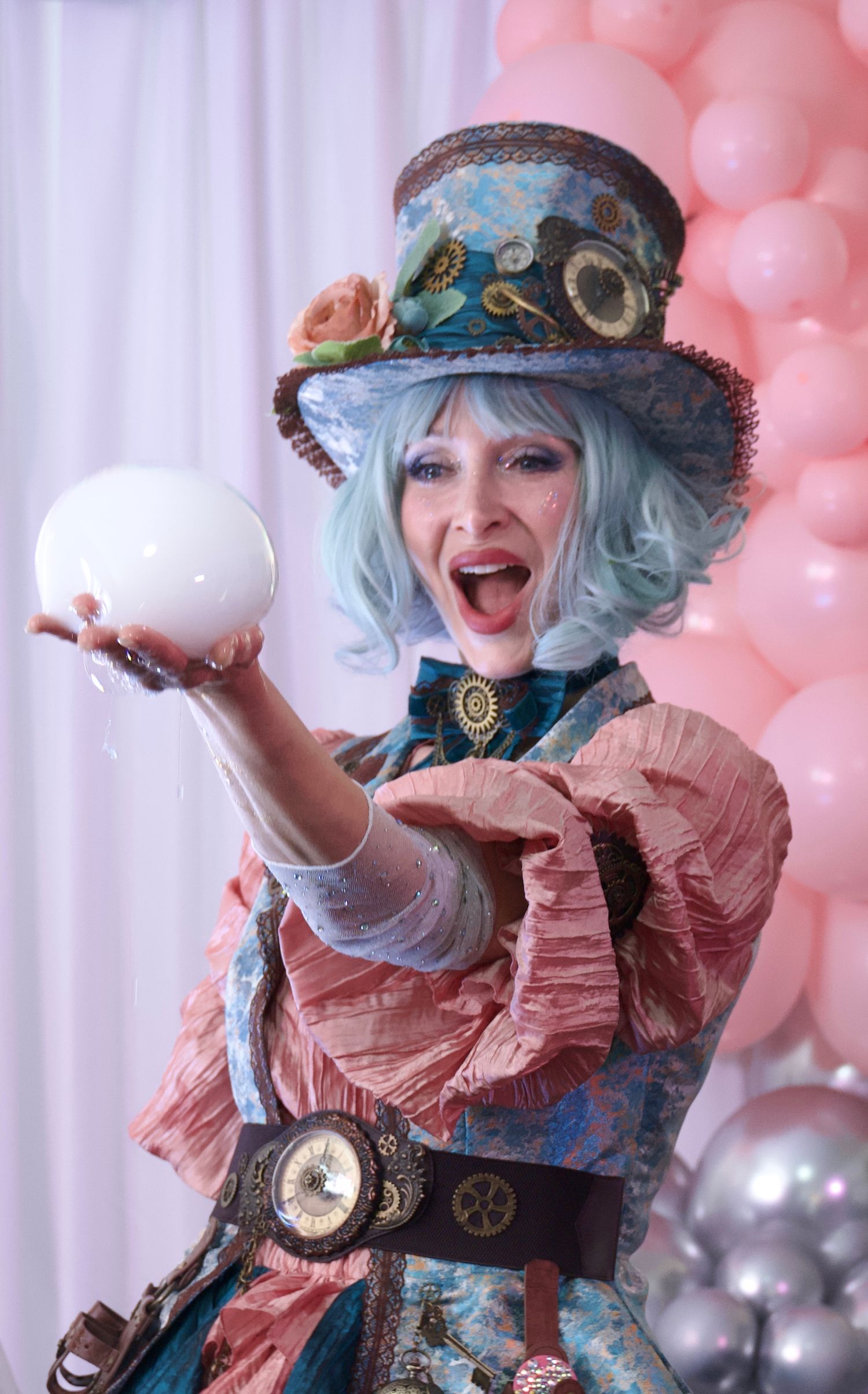 Cosplayer in steampunk attire with blue hair and decorative hat poses playfully with teacups against pink balloon backdrop.