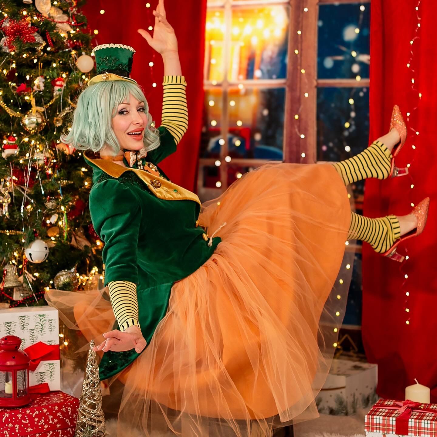 Woman in green top and orange tulle skirt dances playfully by Christmas tree with wrapped presents and festive decor.