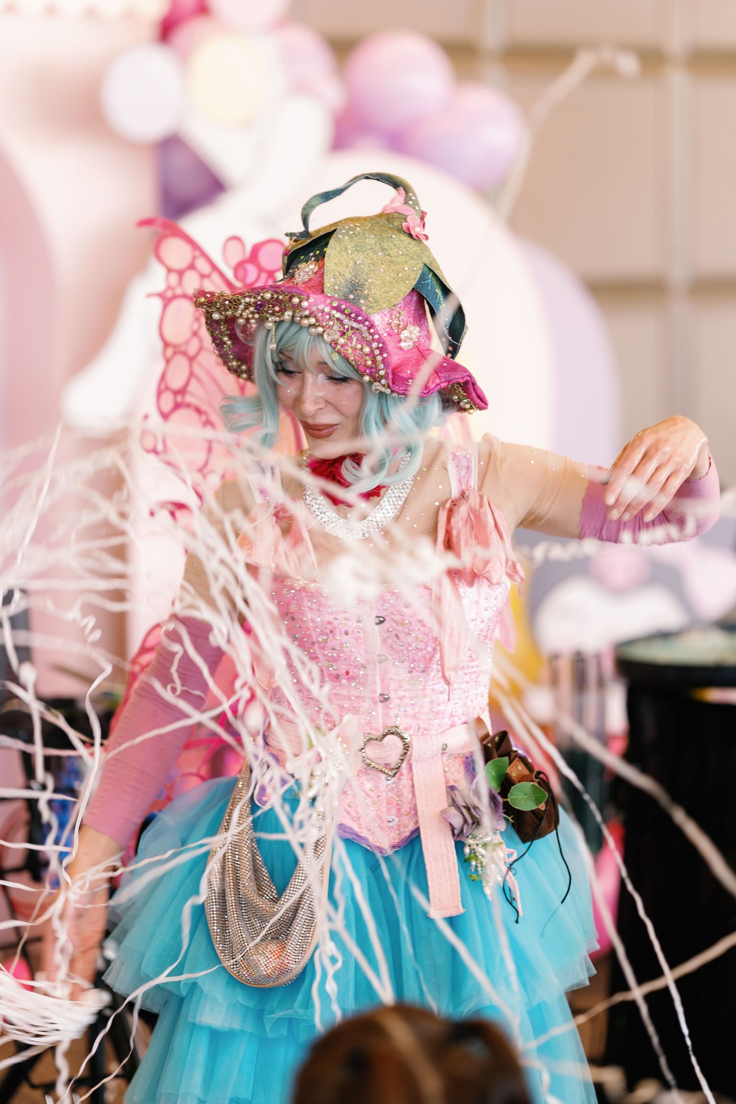 Performer in turquoise and pink outfit surrounded by streaming confetti and decorative elements.