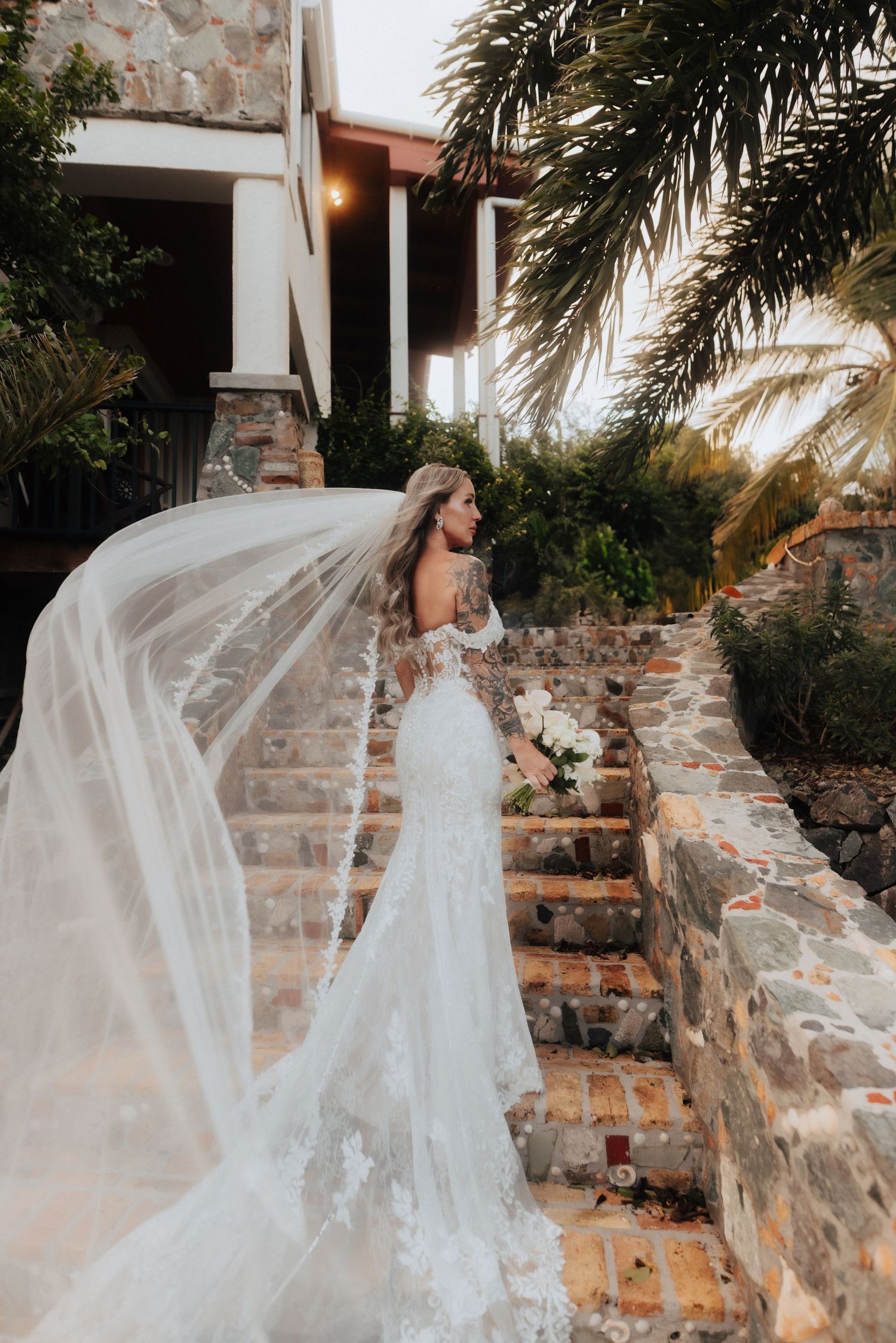 A bride in a lace wedding dress with flowing veil stands on rustic stone steps near palm trees.