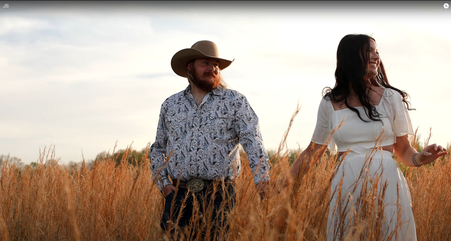 A couple in a wheat field at golden hour, with one wearing a cowboy hat and patterned shirt.