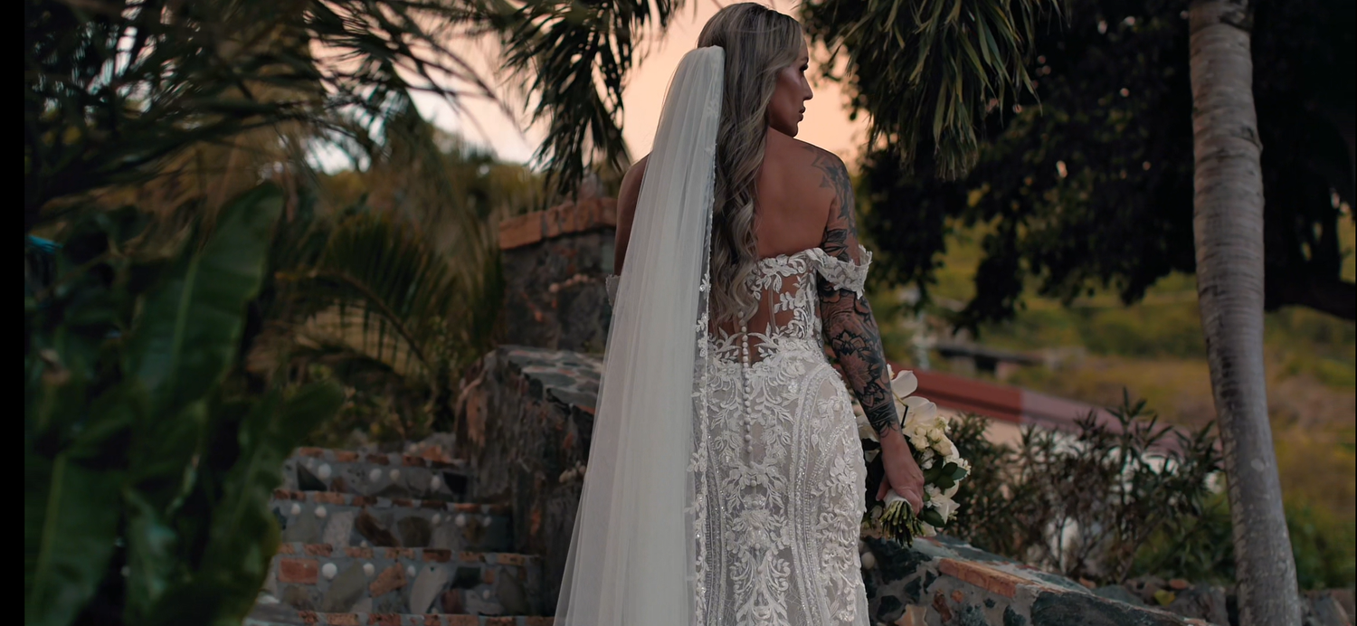 A detailed lace wedding dress silhouetted against a tropical sunset with palm trees.