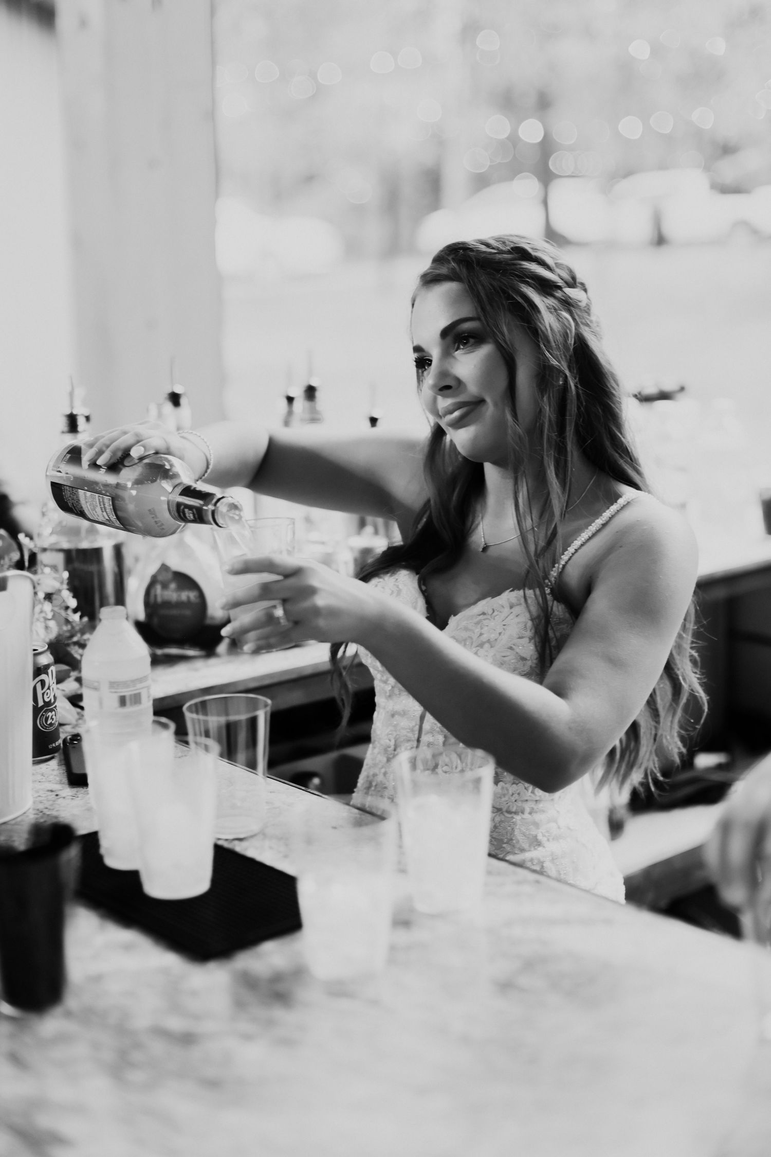 Black and white photo of bride pouring drinks at wedding reception bar.