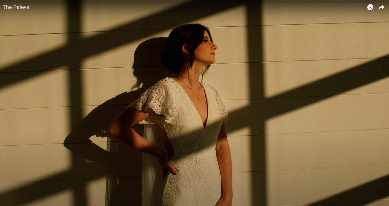 A person in white clothing stands in dramatic shadow patterns against a beige wall.