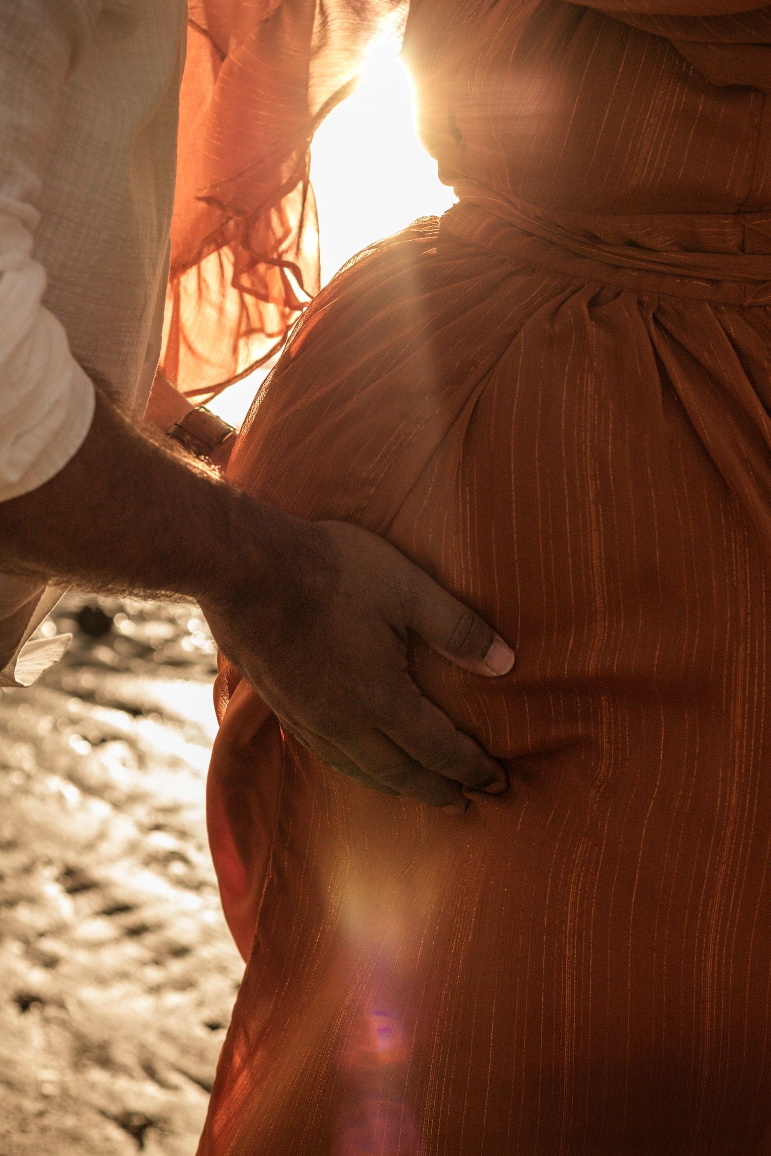 Pregnant mother's belly and father's hands holding at the beach during sunset maternity photography session in Hervey Bay.