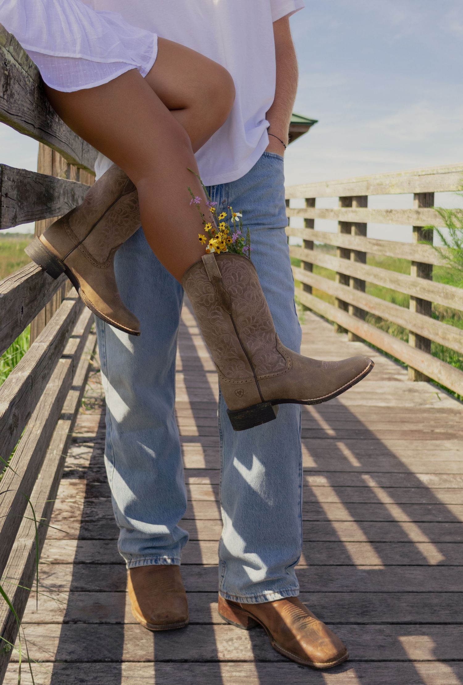 Couple on wooden bridge, woman wearing cowboy boots with wildflowers tucked inside them.