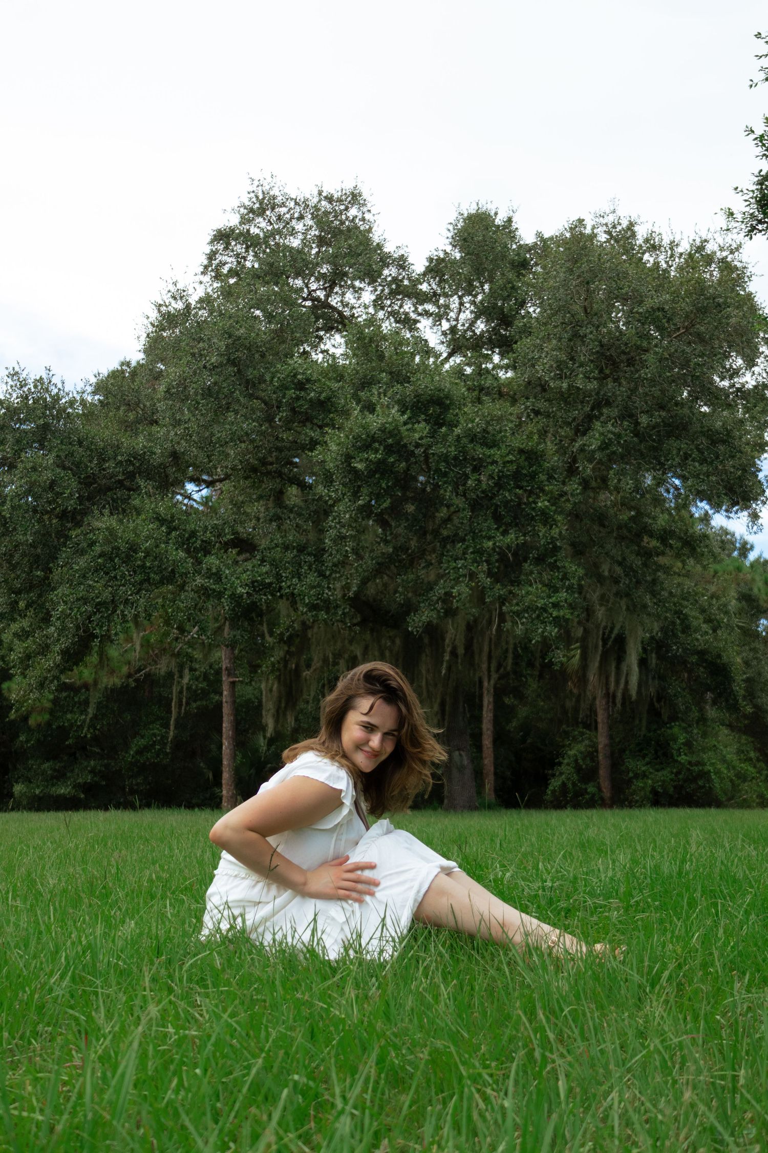 Young woman in white dress sitting and laughing in a vibrant green field with large oak trees.