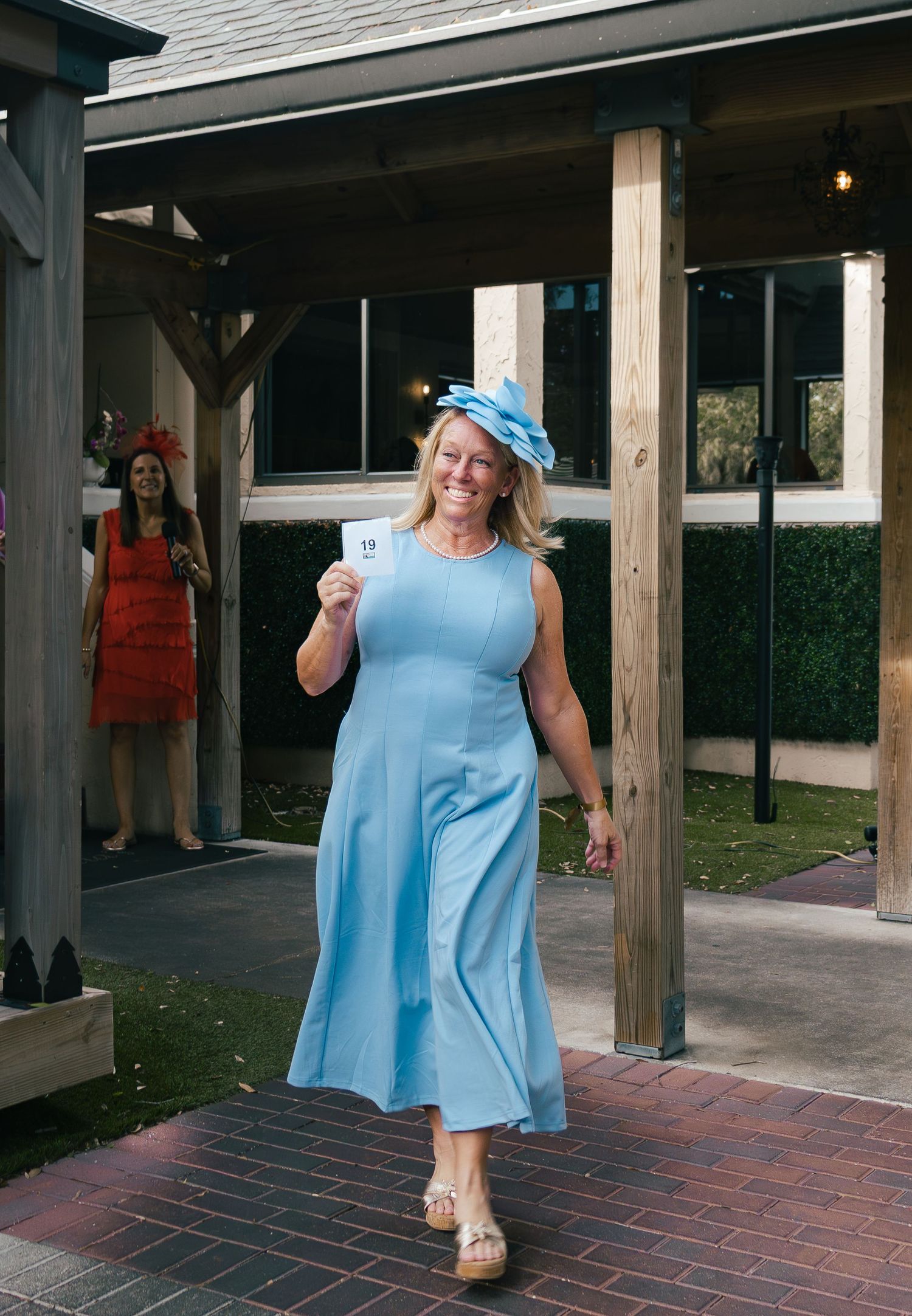 Woman in light blue dress and floral hat holds a card while walking at an outdoor Kentucky Derby event.