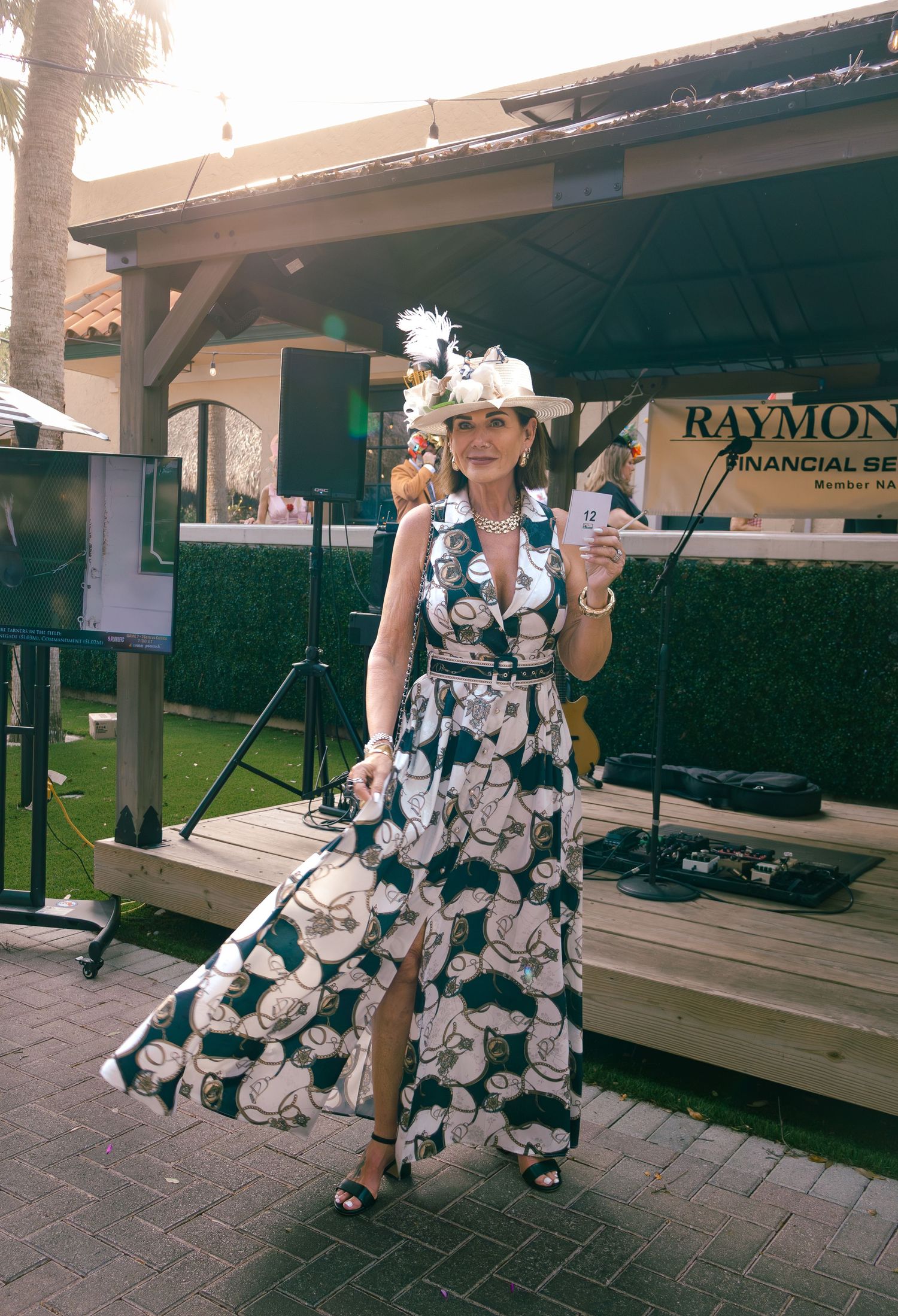 Woman in floral maxi dress and white fascinator hat poses outdoors at a Raymond James Financial Services event.