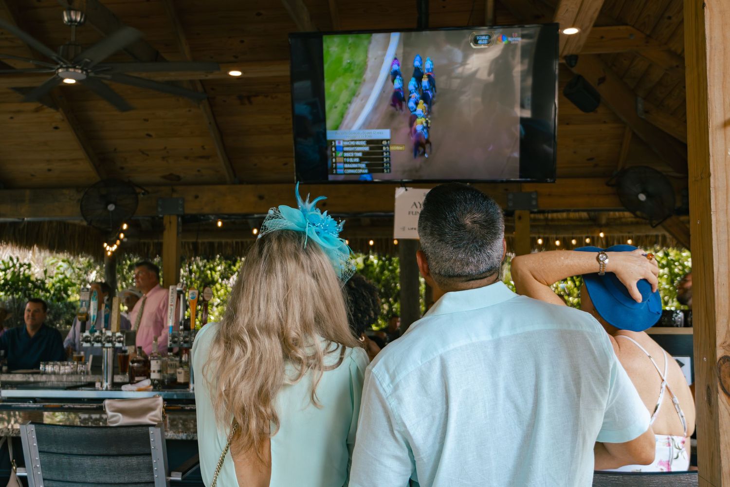 Guests in festive hats watch horse racing on an outdoor TV at a Kentucky Derby party.