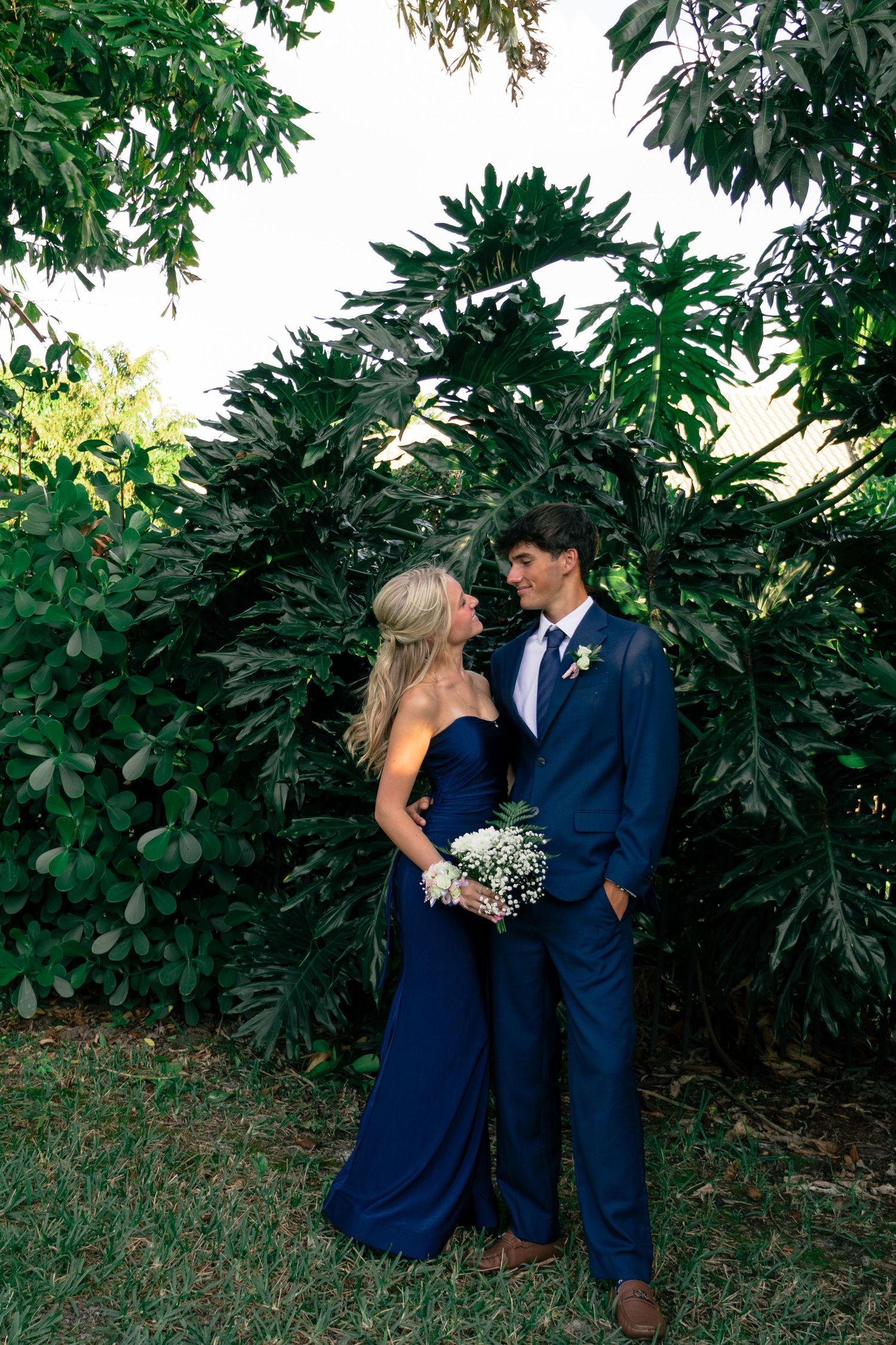 Prom couple in matching navy blue outfits gazes at each other surrounded by tropical green foliage.