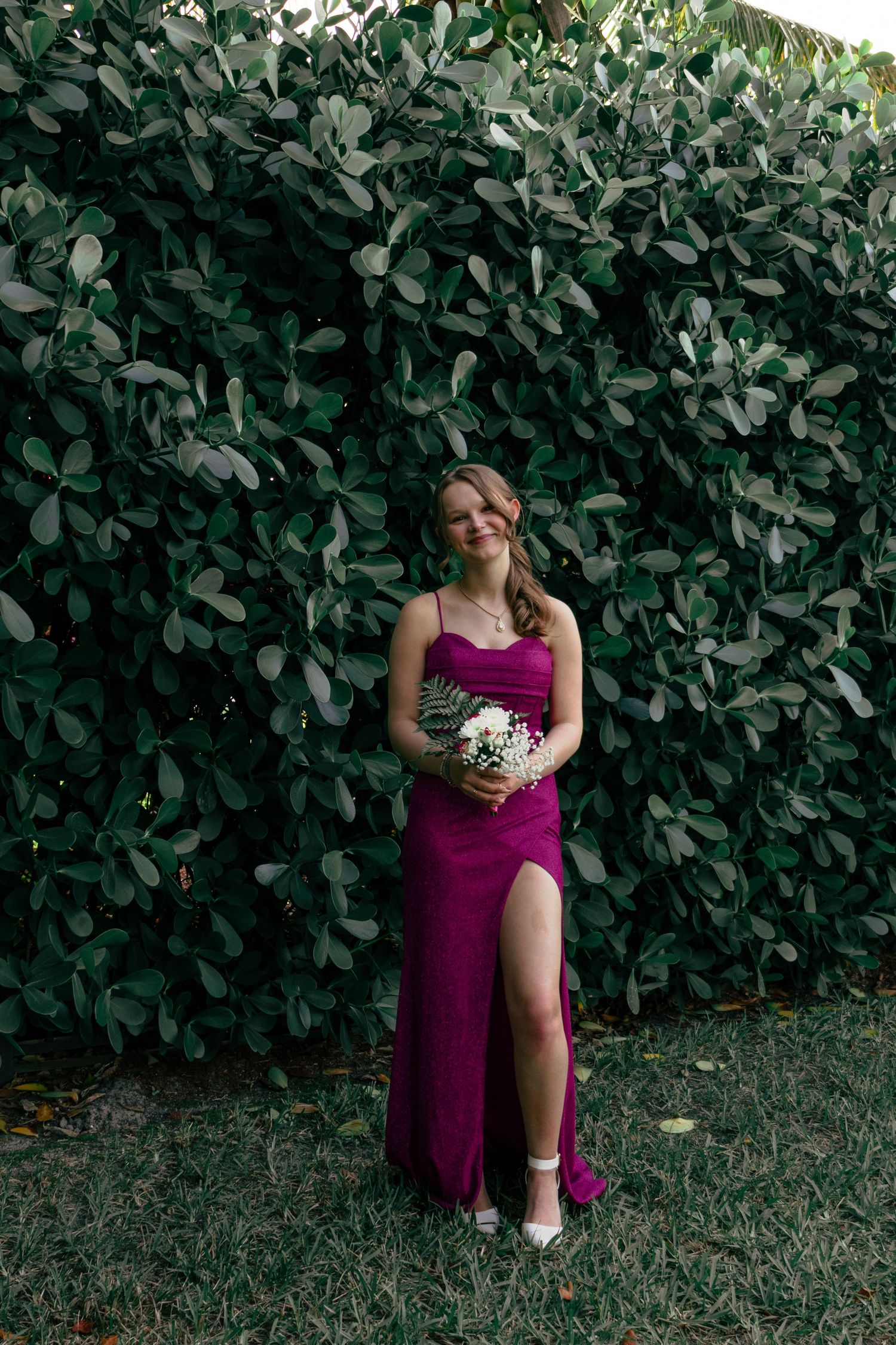 Young woman in magenta slit gown holding bouquet poses before lush green hedge wall.