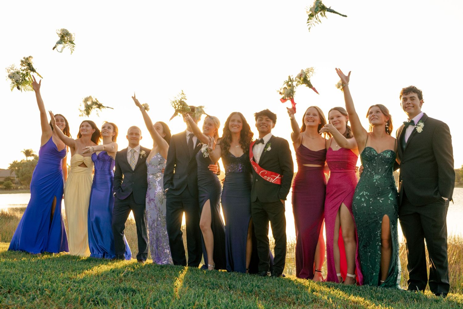 Large prom group celebrating outdoors at golden hour tossing bouquets joyfully.