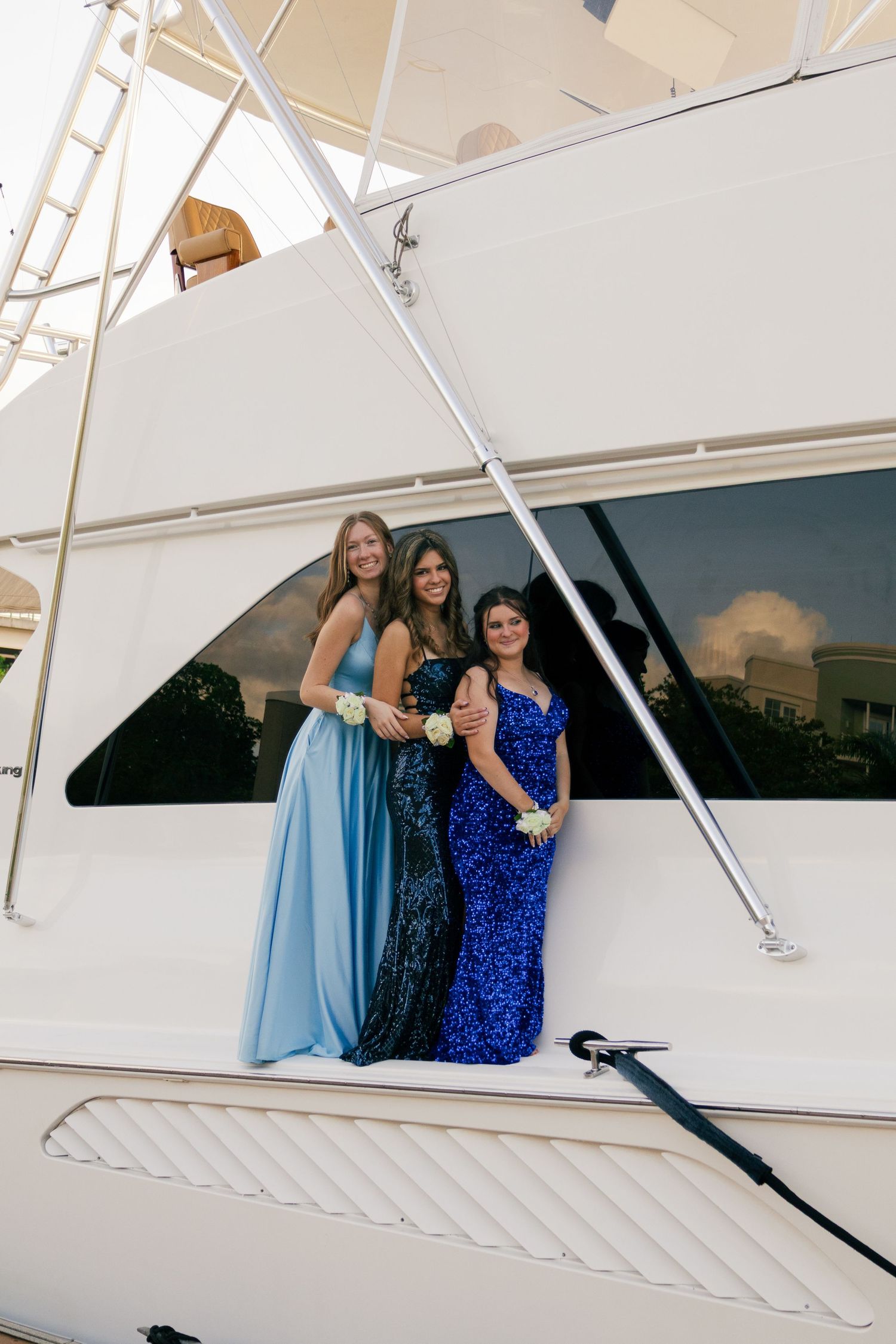 Three teenage girls in blue prom dresses pose together on the deck of a white luxury yacht.
