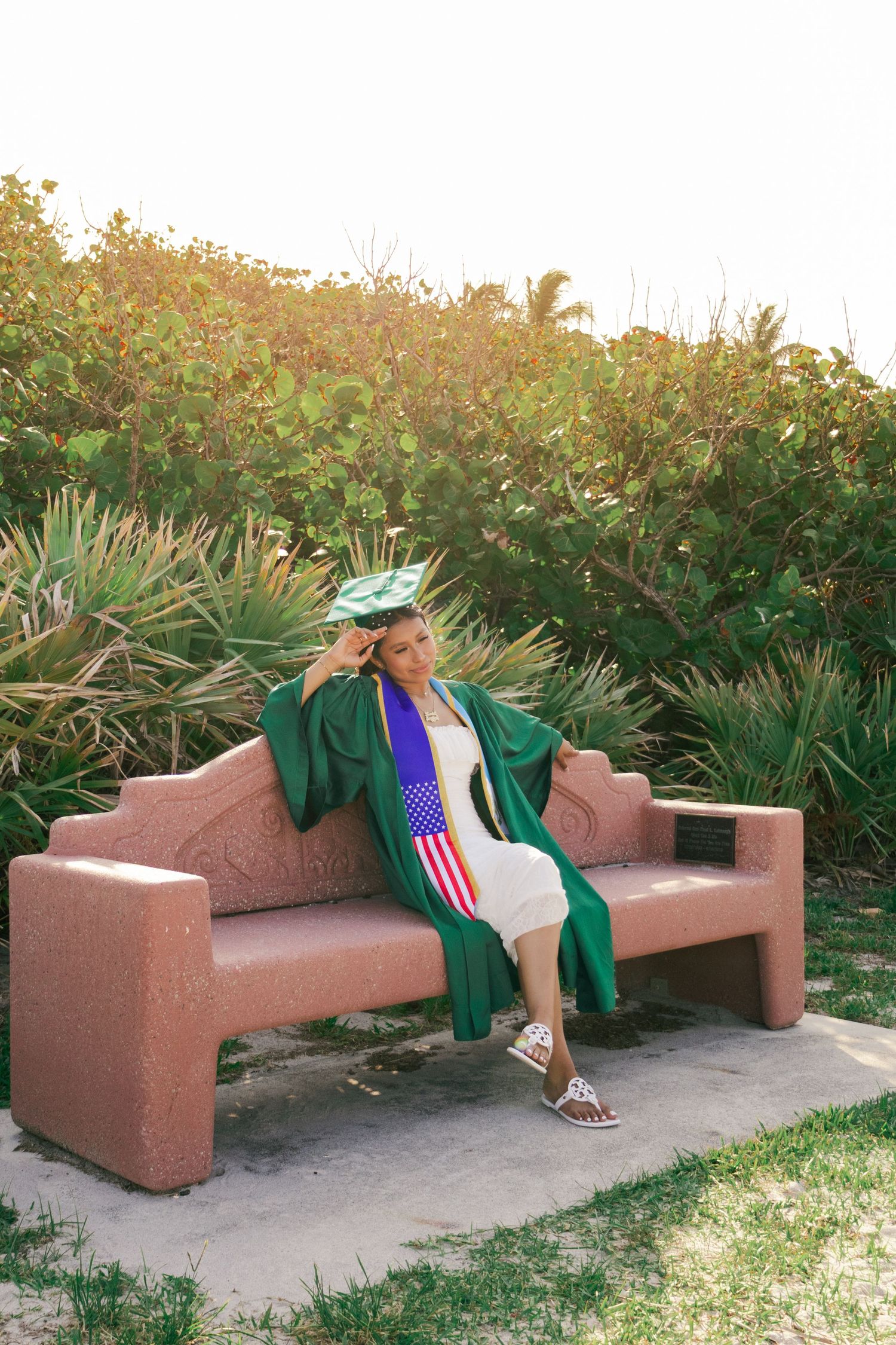 Graduate in green cap and gown with American flag stole relaxes on pink bench in garden.